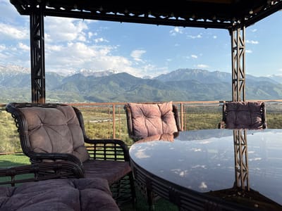The image depicts an outdoor seating area overlooking a mountainous landscape. In the foreground, there are several wicker chairs with plush cushions, and a round glass-topped table. The table reflects the sky, clouds, and a structural support of the covered seating area. Beyond the seating area, a railing overlooks a verdant valley with scattered buildings. In the distance, a range of majestic mountains, some with snow-capped peaks, forms the horizon under a partly cloudy blue sky. The scene suggests a tranquil setting, likely in a resort or a private residence with a scenic view. The location is identified as Ryskulov, Kazakhstan.