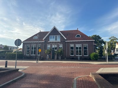 The image shows a brick building with a red tile roof, located at the intersection of Bovenmeester and Sint-Vitushout in Steenwijk, Netherlands. The building appears to be a restaurant or cafe, with signs that read "bovenMeester" and "lunch · diner · lounge". There are large windows on the facade, and several tables and chairs are visible outside, suggesting an outdoor seating area. A bicycle is parked near the entrance, and a yellow sign with Dutch text is posted on a pole. The street in front of the building is paved with red bricks, and there is a crosswalk. The sky is blue with scattered white clouds. The overall scene depicts a bright, daytime setting in a European town.