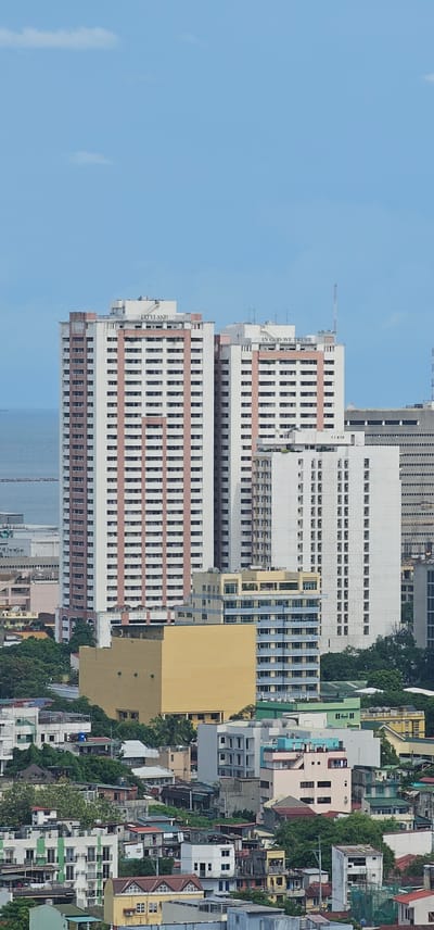 This image captures a high-angle view of a dense urban landscape in Makati, Philippines, under a clear blue sky with scattered white clouds. The scene is dominated by a cluster of high-rise buildings of varying architectural styles and colors, interspersed with lower-rise residential structures and lush green trees.

In the foreground, a collection of multi-story buildings with diverse facades, including some with green and white striped exteriors, are visible. Red-tiled roofs are prominent on some of these structures. Further back, a large, monolithic, yellow building stands out.

The mid-ground is characterized by several white and off-white apartment buildings. The most prominent among these are two tall, cream-colored towers with reddish-brown vertical accents and numerous uniformly spaced windows. The word "CITYLAND" is inscribed in black lettering on the top of the left tower, and "IN GOD WE TRUST" is visible on the top of the right tower. Adjacent to these, another white high-rise with a grid-like pattern of windows is situated.

In the background, a larger, darker building with a visible grid of windows, likely another office or residential building, is partially obscured by the closer structures. The distant horizon shows a glimpse of the sea, suggesting a coastal location within the city. The overall impression is a densely populated urban environment with a mix of modern skyscrapers and older, more traditional housing. No people are visible in the image.