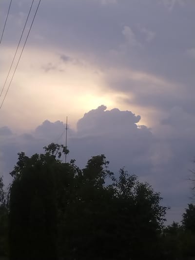 The image depicts a sky with dramatic clouds and the sun peeking through. In the foreground, silhouetted trees and a tall antenna with wires are visible. The scene suggests an outdoor environment in Ivatsevichy, Belarus, likely during late afternoon or early evening given the lighting conditions. There are no people or discernible actions taking place. The overall atmosphere is somewhat moody due to the heavy cloud cover and the dim lighting.