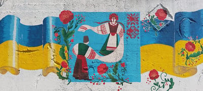 This image depicts a mural painted on a textured, white wall. The mural features a vibrant design that appears to incorporate elements of the Ukrainian flag, with broad bands of blue and yellow dominating the lower portion of the composition. The blue band is on top, followed by the yellow band. These bands are depicted as if they are draped or folded, creating a dynamic background.
Central to the mural are two figures painted in a folk art style. The figure on the right is depicted in profile, with a white headdress and long braided hair, wearing a traditional white garment adorned with red embroidered patterns on the sleeves and neckline, and a dark brown vest over a green dress. The figure on the left, also in profile and facing the other figure, wears a dark brown conical hat, a green tunic, and a red skirt. Both figures are positioned within a central blue rectangular area, surrounded by stylized red floral motifs and green branches with red berries.
To the left and right of the central blue area, the blue and yellow bands of the flag continue, interspersed with painted red flowers, resembling poppies, and green foliage. The flowers and branches are rendered in a decorative, folk style with bold outlines and bright colors. The white background wall shows signs of wear and texture, with some peeling paint visible. The mural appears to be located outdoors. The provided context states this mural is located in the city of Cherkasy, Ukraine.