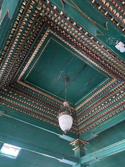 The image shows a close-up, low-angle view of a highly ornate, green painted ceiling with extensive gold and red decorative elements. The ceiling is tiered, with each layer adorned with intricate patterns. The central area of the ceiling is flat and green, with a decorative molding framing it. Suspended from the center of the ceiling is an antique-looking white glass pendant lamp, hanging from a dark metal chain. The surrounding areas of the ceiling feature multiple rows of repeating geometric patterns, alternating between red and gold leaf accents. Along the edges of the ceiling, there are carved green decorative elements that extend downwards. On the upper right of the image, a white light fixture is visible, mounted on the ceiling. A small square window with bright light emanating from it is visible in the lower left corner. This ceiling is located within the Kasepuhan Palace (Keraton Kasepuhan) in Cirebon, Indonesia, specifically in the Jenggala room, which is known for its unique architecture and historical significance.