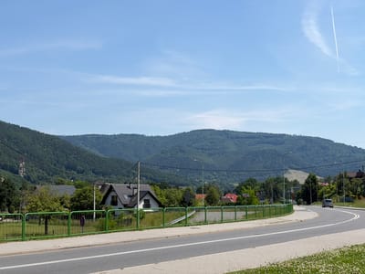The image depicts a scene in Międzybrodzie Bialskie, Poland, on a clear, sunny day. In the foreground, a two-lane asphalt road curves to the right, bordered by a concrete sidewalk and a grassy area with white wildflowers. A green metal fence runs along the edge of the sidewalk.
Beyond the fence, a residential area is visible with several houses featuring dark-tiled roofs. One house, predominantly white with dark trim, is prominent. Trees and vegetation are scattered throughout the settlement. In the background, a lush, tree-covered mountain range dominates the landscape. The sky is a bright blue with a few wispy white clouds and a long contrail from an aircraft.
A silver car is captured in motion on the road, heading away from the viewer and around the curve. Electrical poles and wires are present, particularly on the left side of the image, and a radio tower is visible in the distance on the left. The overall impression is a peaceful, rural or suburban setting nestled against a natural mountainous backdrop.