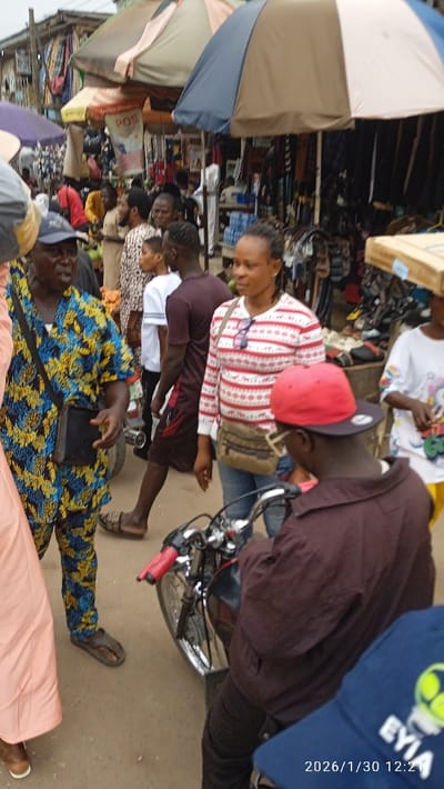 The image depicts a crowded outdoor street market in Ijaiye, Nigeria. The environment features an unpaved or dirt ground surface and multiple large market umbrellas of varying colors (blue, white, tan, light tan) providing shade over numerous stalls. These stalls display various goods, including clothing, bags, and general merchandise; a "POS" (Point of Sale) sign is visible on a banner in the background.
In the mid-ground left, an older male with dark skin wears a blue and yellow patterned short-sleeved outfit, brown flip-flops, and a dark cap. He carries a black shoulder bag and is oriented towards the left. Adjacent to him, towards the center-right, a female with dark skin and braided hair wears a long-sleeved white shirt with red horizontal stripes, blue denim jeans, and brown flip-flops. She has sunglasses tucked into her shirt collar and carries a tan cross-body bag, looking slightly to her left.
In the foreground right, a person with dark skin, seen from the back, wears a dark brown long-sleeved shirt or jacket and a red baseball cap. This individual is seated on a motorcycle with a dark fuel tank, chrome components, and red handlebar grips. A blue cap with a logo and "EYIA" text, along with a date stamp "2026/1/30 12:21," is visible in the lower right corner. Numerous other individuals populate the background, walking and standing among the market stalls.