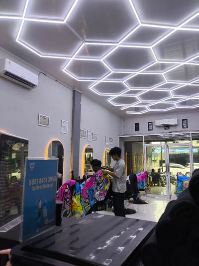 The image depicts the interior of a barber shop located in Miruektaman, Indonesia. The shop features a modern design with a hexagonal LED lighting system on the ceiling and recessed arch lighting behind mirrors. An air conditioning unit is mounted on the wall. In the foreground, a barber is attending to a client, using clippers. The client is seated, wearing a vibrantly patterned cape. To the left, a blue sign displays a phone number "0851 8451 3953" and the name "Syibrall Malasyi". Reflections in a mirror on the left show the exterior of the shop, including a white car. Through the glass doors at the front, other patrons can be seen sitting and interacting, with motorcycles parked outside. The overall atmosphere suggests a contemporary and well-lit establishment.