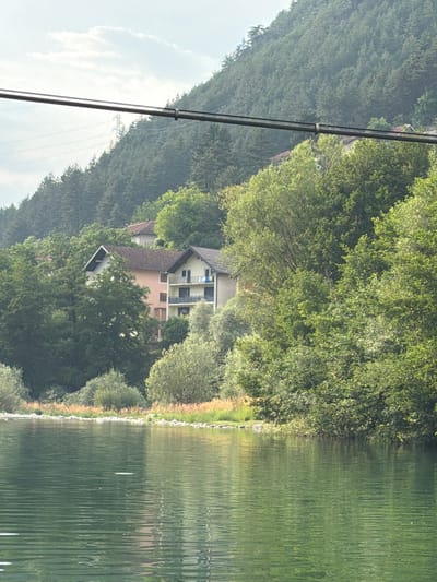In Konjic, Bosnia and Herzegovina, a serene lake reflects the lush green trees and a multi-story building nestled on a hillside. A transmission tower is visible in the distance, perched atop a forested mountain. Power lines crisscross the sky, creating a stark contrast against the natural landscape. The water's surface is rippled, suggesting a gentle breeze. The scene evokes a sense of tranquility and highlights the harmonious coexistence of nature and human infrastructure.
