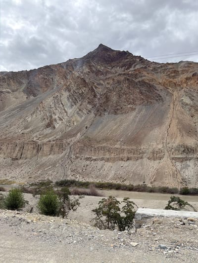 The image shows a mountainous landscape in the city of Chilling, India. The dominant feature is a large, barren mountain with distinct geological layers and striations, colored in shades of brown, beige, and gray. The mountain has a sharp peak. In the midground, a brown river flows through a wide, dry riverbed lined with sparse vegetation and small trees. The foreground consists of a gravelly, dusty terrain with scattered rocks and some more vegetation. The sky is overcast with gray clouds. There are no people or animals visible in the scene. Several utility wires are strung across the upper part of the mountain.