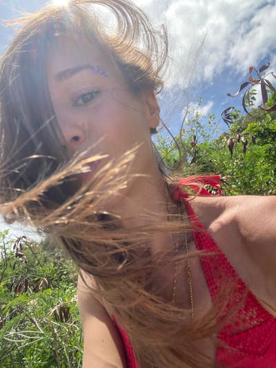 A low-angle, close-up photograph captures a person with light skin, brown hair, and a possible blue eyebrow decoration against a background of sky, trees, and vegetation. The person is wearing a red top and gold necklaces. The location appears to be in a natural environment, potentially a park or garden. There is no specific information within the image to confirm this is Baan Kai, Thailand.