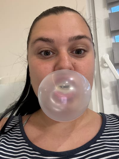 The image shows a close-up, frontal view of a woman in Rogosh, Bulgaria, blowing a large bubble gum bubble. The woman has dark hair pulled back, prominent eyebrows, and is wearing a dark and white striped tank top. Her eyes are wide open, and her mouth is pursed around the bubble. The bubble itself is translucent, reflecting light and obscuring her lips and chin. In the background, a white window handle and blinds are visible.