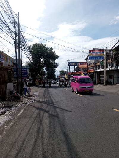 This image captures a wide-angle view of a bustling urban street scene in Domphoog_Dungkarchhoeling, Bhutan, under a partially cloudy daytime sky.
In the immediate foreground on the left, an unpaved shoulder with dark patches of moisture or grime abuts the asphalt road. A dark utility pole with a dense network of black electrical and communication wires dominates the left side, casting long, distinct shadows diagonally across the road towards the right. Beneath these wires, a male individual stands on the roadside, wearing a white cap, white short-sleeved shirt, and blue jeans, appearing to interact with a small, light-colored object or cart at his feet. Behind him, a commercial building features a partial sign with the text "PANEN PROMO" and other Indonesian script.
The asphalt road, marked with a solid yellow line on its right edge, shows moderate traffic. A bright pink Mitsubishi Colt L300 minivan, with white hubcaps, is prominently positioned in the right lane, facing away from the viewer. Further down the road, approximately five to six motorcycles, ridden by individuals, are visible, largely traveling in the same direction as the minivan. Additional distant vehicles, including cars and other vans, are present in the far background.
A large, dense green tree stands centrally in the midground, its foliage contrasting with the overhead wires. On the right side of the street, commercial buildings line the thoroughfare, featuring prominent advertising signs. Visible signs include "Indomaret" (a red and blue logo), "Alfamart" (a red and white logo), and a blue sign reading "BPR K...". The overall environment suggests an active commercial district with a mix of vehicle traffic and pedestrian presence.
