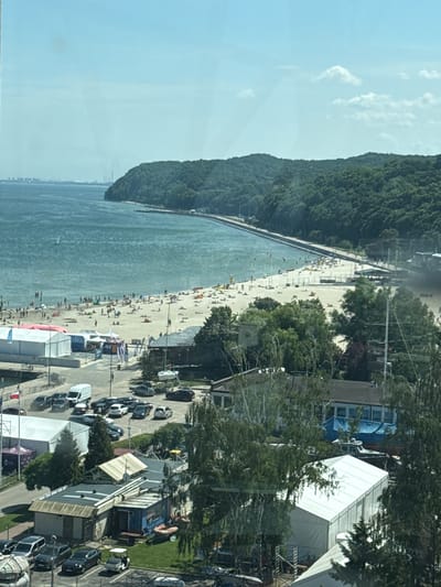 The image shows a crowded beach on a sunny day, with a body of water stretching into the distance. A dense, green forest covers a hillside that runs along the edge of the beach. Several people are scattered across the sandy shore, some sunbathing, others walking or playing. A road runs parallel to the beach, with numerous cars parked along it. Structures, including white tents and buildings, are visible behind the road, suggesting a developed area or event taking place. The sky is bright and clear, with a few wispy clouds, indicating fair weather. The overall scene suggests a popular tourist destination during peak season.