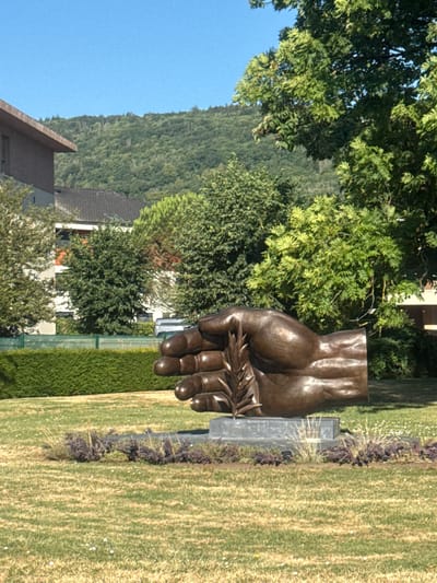 The image depicts a bronze sculpture of a cupped hand holding a single stalk of wheat, positioned outdoors on a sunny day. The sculpture is the central focus, situated on a grey stone base surrounded by low-lying, dark purple foliage and ornamental grasses. Behind the sculpture, a neatly trimmed green hedge forms a boundary, and beyond that, the facade of a modern building with a brown roof is visible. Further in the background, trees with lush green foliage and a distant, tree-covered hillside under a clear blue sky complete the scene. This sculpture is located in Divonne-les-Bains, France, specifically in the Jardin des Thermes.