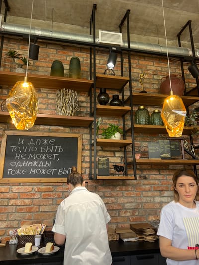The image, oriented as a portrait, captures a café setting with a rustic brick wall as the backdrop. The main subjects are two women, one in the foreground and another partially visible, both wearing white tops. The woman in the foreground, facing away from the camera, is wearing a white shirt and is bending down towards a counter where two slices of cheesecake are displayed on small plates. She has her hair tied up in a bun and is wearing earrings. The second woman, positioned in the upper right of the frame, is looking towards the camera and wearing a white t-shirt with a subtle striped pattern.
The scene is filled with various objects, including decorative lamps with a golden hue, green vases, and shelves stocked with books, plants, and decorative items. A large chalkboard with white lettering is prominently displayed on the right side of the image, featuring what appears to be a menu or a quote in Cyrillic script. The text on the chalkboard reads: "и даже то, что быть не может, однажды тоже может быть". Some other text in smaller font on a separate board is also visible. The overall ambiance suggests a cozy and perhaps artisanal establishment, possibly a café or a small eatery, given the presence of food items and menu boards. The lighting is warm, hinting at an indoor setting, possibly during daytime.