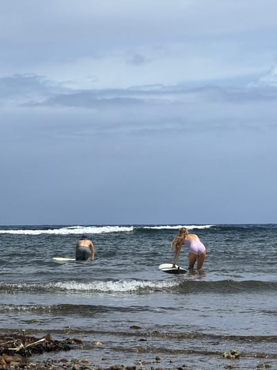 A vertically oriented image displays a beach scene. The left half features a cloudy sky. The right half shows ocean water meeting a shore. A woman in a pale pink bodysuit bends over a surfboard in the water. Further along the shoreline, a man with a blue cap is waist-deep in the water with a paddle. The beach is rocky and uneven. The location is assumed to be Santa Monica, Philippines.