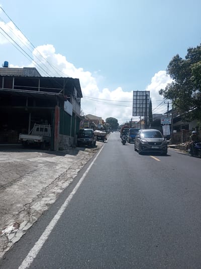 The image depicts a street scene in an unidentified urban area, not demonstrably within Zeyathiri District, Myanmar. The road is asphalt and has a single white line marking the shoulder. Several vehicles are present, including a gray car in the foreground, other cars and small trucks further down the road, and a motorcycle. Buildings line the left side of the street, with some appearing to be commercial establishments with open-front garages or storage areas. A billboard is visible in the distance above the vehicles. The sky is blue with scattered clouds. Vegetation, including trees, is present on the right side of the street. The overall lighting suggests a daytime setting. There is an absence of explicitly discernible activity or interactions in the frame.
