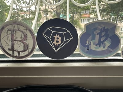 Three stickers are placed on a windowsill, with a window and foliage visible in the background. The stickers appear to be related to cryptocurrency. The leftmost sticker is circular and features a metallic-looking "B" symbol with lines radiating from it, and the word "Bitcoin" is visible at the bottom. The middle sticker is also circular, with a dark background and a white outline of a diamond shape containing a "B" symbol. The rightmost sticker is die-cut and depicts a stylized 3D "B" symbol within a geometric design, with small figures of people around it. The windowsill is white and shows some dust and grime. The environment outside the window suggests an urban setting with buildings and trees. The image was taken in Nakhodka, Russia.