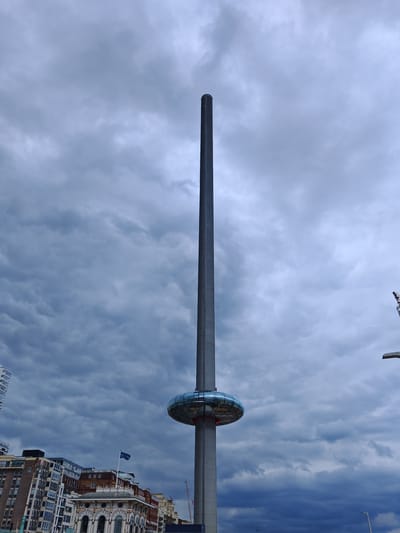 The image shows the British Airways i360 observation tower in Brighton, United Kingdom, under a cloudy sky. The tower is a tall, slender, gray structure with a circular, glass-enclosed observation pod located approximately one-third of the way up. The sky is filled with gray clouds, suggesting overcast conditions. Buildings are visible at the bottom of the frame, indicating an urban environment. The location is specifically in Brighton, which is not within the borough of Reigate and Banstead. There are no people or specific interactions visible in the image.