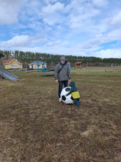 The image, taken in Ludorvai, Russia, depicts an outdoor scene on a partly cloudy day. The setting is a large, open grassy field, characterized by sparse brown and green patches, suggesting early spring or late autumn.
In the foreground, an adult male and a small child are featured with an oversized white and black soccer ball. The adult, dressed in a grey beanie, a dark olive-green jacket, and dark trousers, stands beside the giant ball, looking towards the camera with a gentle smile. He has a dark shoulder strap across his jacket. The child, wearing a dark green or blue full-body suit with a bright yellow upper portion, is playfully leaning or lying on top of the large soccer ball, appearing to interact with it.
The background reveals a rural landscape. Several houses are visible in the middle ground, including a distinctive yellow house with white windows. A blue-framed trampoline is also present among the houses. Behind these structures, a dense line of pine trees stretches across the horizon, indicating a bordering forest. In the lower-left foreground, a grey slide or ramp is partially visible. The sky above is bright, with scattered white and grey clouds against a clear blue expanse. The overall scene conveys a relaxed and playful daytime moment between the adult and child. No visible text is present in the image.