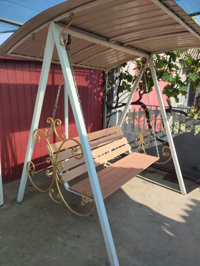 A three-seater swing set with a metal frame and a fabric canopy is positioned outdoors on a concrete surface. The swing set is painted white and has decorative gold metalwork on the sides of the seat. The seat is made of wooden planks painted a light brown color. Behind the swing set is a red corrugated metal fence, and to the right, there is a gray fence with a dark green vine growing over it. Sunlight casts shadows on the ground. The scene appears to be in the village of Komyshivka, Ukraine.