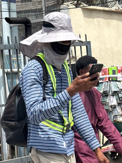 In Falomo, Nigeria, the image captures two men. One, wearing a blue and white striped long-sleeved shirt and a grey sun hat, is seated, using a black mobile phone. He has a neon yellow safety harness over his shirt. The second man, behind him, wears a maroon jacket and is also using a mobile phone. Behind them, stacks of what appear to be phone cases are visible. They are sitting on a metal structure of some kind. The background includes a wall and some overhead cables.