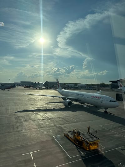 A China Eastern Airlines aircraft is parked on a paved airport apron, casting a long shadow in the afternoon sun. Next to it is a yellow cargo handling vehicle. The sky above is blue with wispy clouds. The image appears to be taken from inside a building or airport terminal, due to the reflections on the glass. The location is likely an airport in Исаково (луневское С/п, Russia), given the presence of China Eastern Airlines, suggesting international travel. No people are visible.