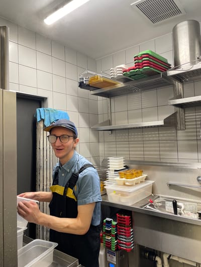 A person wearing a blue button-down shirt, black overalls with a yellow bib, a baseball cap, and glasses stands in a kitchen and holds up a clear plastic container. The person is looking at the camera and smiling. The kitchen has tiled walls and stainless steel shelves that hold stacked red and green plastic containers, along with a large metal pot. There are also stacks of white containers and a tray of small yellow containers on a counter. A sink is visible to the right, and a black and silver appliance is on the floor.