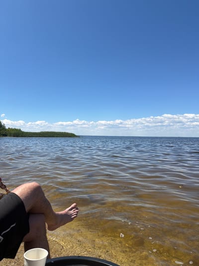 The image depicts a serene lakeside scene under a bright blue sky with scattered white clouds. In the foreground, a person's legs, clad in black shorts, are visible, with one bare foot resting in the shallow, clear water. The water's surface ripples gently, revealing the sandy bottom beneath. The shoreline is lined with green reeds or tall grass, with a dense line of trees further in the distance on the left side of the frame. The expansive body of water stretches towards the horizon, where it meets the sky. The overall impression is one of relaxation and natural beauty, possibly at a beach or recreational area near Lebedivka, Ukraine.