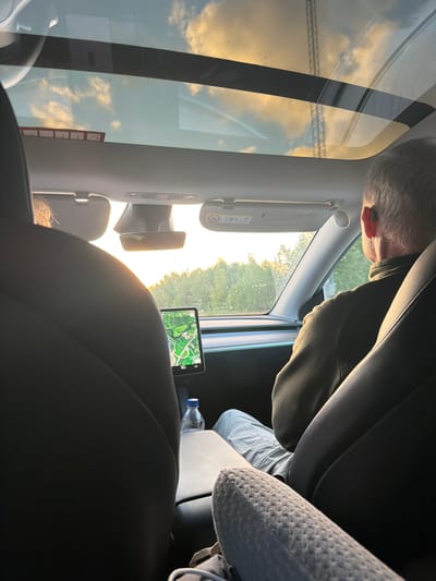 The image captures the interior of a car, likely a Tesla, with a panoramic glass roof showing a cloudy sky with hints of sunset colors. The view extends to the outside, revealing a treeline and what appears to be a body of water. In the foreground, two car seats are visible. In the driver's seat, a man with graying hair is seen from the side, wearing a dark jacket and light-colored pants. He has an earpiece in his right ear. In the passenger seat, a person's back is visible, with blonde hair peeking out. The car's touchscreen display is showing a map. A bottle of water sits on the center console. The image appears to be taken on a road, possibly on the E6 highway near Munkedal, Sweden, given the visible landscape and road signs indicating location.