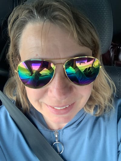 A woman with blonde hair and sunglasses is taking a selfie in a car. She is wearing a blue hooded jacket with a zipper. The sunglasses reflect the dashboard and the road ahead. A seatbelt is visible across her chest. The background shows a car seat. The image appears to be taken in the city of Azerski, Belarus.