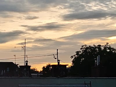 The image shows a view of rooftops and a cloudy sky at sunset. Silhouetted against the orange and yellow hues of the sky are several structures including chimneys and antennas. Trees are visible on the right side of the frame. The overall scene suggests an urban setting in Mława, Poland.