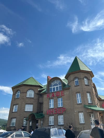 The image shows a multi-story stone building with a green roof, identified by red lettering as "КАФЕ ИНЗЕР" (Cafe Inzer). The building features several windows, some arched, and is situated in a landscape with a bright blue sky and scattered white clouds. In the foreground, several cars are parked, and a few individuals are visible near the entrance of the cafe, as well as walking on the sidewalk. The overall scene depicts a commercial establishment in the town of Inzer, Russia.