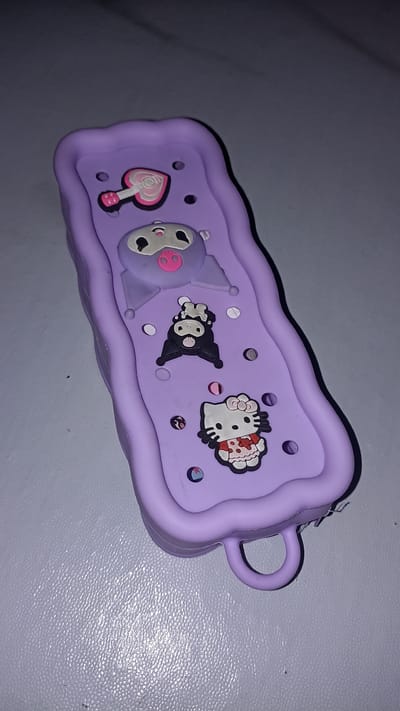 A lavender-colored pencil case with decorative charms sits on a textured surface. The charms include a pink guitar, a grey and pink pig-like character, a black and white cartoon character, and Hello Kitty wearing a red and white patterned dress. The pencil case has a wavy edge and a loop handle. The scene is dimly lit, with shadows casting across the surface. The location is not specified in the image.
