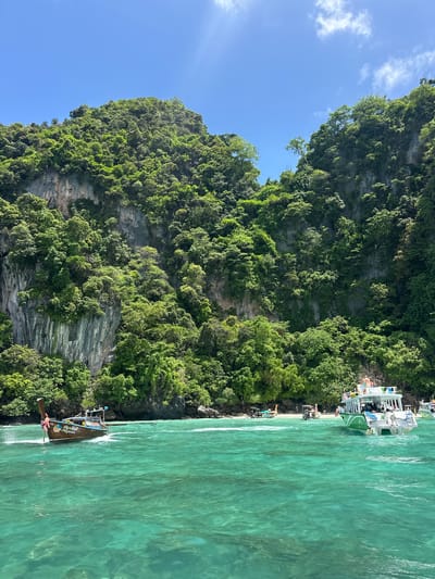 A wide shot captures the turquoise waters and lush, green cliffs of Ton Sai, Thailand. In the foreground, a traditional longtail boat with a person at the helm cuts through the water, leaving a white wake. To the right, a larger white and green tour boat is moored, with several people visible on its deck. In the distance, a small beach is visible, dotted with more longtail boats and a few people. The cliffs are densely covered with vegetation, and the sky above is a clear blue with a few wispy clouds. The overall scene depicts a vibrant tropical paradise.