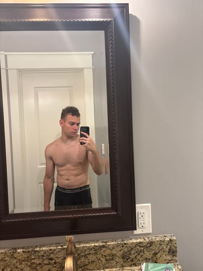 A young man with curly brown hair and a light complexion is taking a mirror selfie in a bathroom. He is shirtless and wearing black boxer briefs. He has a thin gold chain around his neck and is holding a black smartphone in his right hand. He appears to be in his early 20s. The bathroom has a granite countertop, a gold faucet, and a dark brown framed mirror. The walls are painted a light gray. Behind him, a white door is visible. The image was taken in a private residence in Panama City Beach, Florida, United States of America.