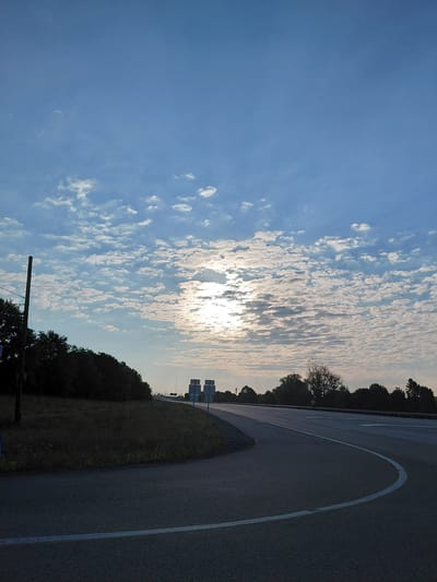The image shows a highway scene during the day in Richmond, United States of America. The sky is partly cloudy with the sun visible through the clouds, creating a bright halo effect. A paved road with white lane markings curves through the scene. On the left side of the road, there is a grassy embankment with trees and a utility pole. On the right side, more trees and road signs are visible in the distance. The overall impression is of a quiet, open highway.