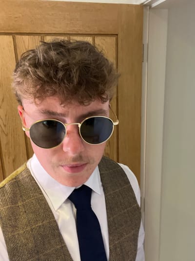 A close-up portrait of a single male individual is depicted in an indoor environment. The subject, an adult, has short, curly brown hair and a light brown mustache. He is wearing gold-rimmed, oval-shaped sunglasses with dark, reflective lenses, obscuring his eyes. His attire includes a white collared dress shirt, a dark blue tie, and a brown and tan checkered or herringbone patterned waistcoat. The individual is positioned facing the camera, with a direct gaze. The background consists of a light-colored wooden door with vertical paneling on the left side of the frame, and a plain light-colored wall with a visible door frame and a metallic hinge on the right. The lighting is consistent with an interior setting. There are no visual cues present within the image to definitively identify its specific location within the city of Hornsea, United Kingdom; the scene appears to be a private or domestic interior.