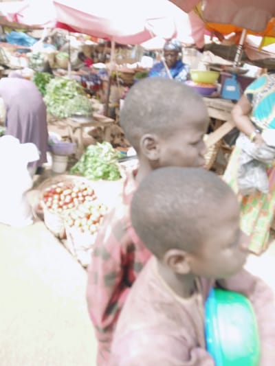 This image captures a vibrant open-air market scene in Kwekwe, Zimbabwe. In the immediate foreground, two young, dark-skinned children are positioned facing right. The child closer to the viewer wears a light brown long-sleeved top and holds a green and blue plastic container against their chest. Behind this child, another young individual wears a red and black checkered long-sleeved shirt. The background, though partially out of focus, reveals several market stalls shaded by various large umbrellas, including pink, orange, and white. Piles of fresh green leafy vegetables are prominently displayed on low surfaces and the ground. Bags containing what appear to be small red fruits or nuts are also visible. Multiple adults populate the market; one person in a purple garment has their back to the viewer on the left, while another adult in a blue headscarf is positioned behind a stall with a blue weighing scale and yellow bowls. An individual wearing a patterned dress is partially visible on the right. The ground appears to be unpaved or dirt.