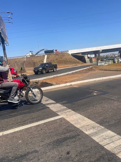 The scene depicts an outdoor urban environment in Anápolis, Brazil, under a clear blue sky. In the foreground, a person wearing a gray long-sleeved shirt over a maroon garment, gray pants, and gray sneakers is seated on a red motorcycle. The person's right hand holds a red smartphone. The motorcycle is positioned on a paved road marked with white perpendicular lines, indicative of a crosswalk.

In the midground, a dark-colored Ford Ranger pickup truck is driving from left to right on an ascending asphalt road section. Above this, on an elevated roadway, road construction is underway. A white asphalt milling machine is actively operating, feeding material into the open bed of a red dump truck. This elevated roadway is part of a concrete overpass structure, supported by multiple pillars, which spans across the lower roads.

To the right, near the overpass pillars, several commercial billboards and signs are visible, some partially obscured. In the far background, a dense cluster of urban buildings can be seen under the clear sky. Grassy and dirt embankments flank the elevated road. On the far left, a utility pole supports a large, unreadable billboard structure. The overall lighting suggests direct sunlight, casting shadows across the scene.