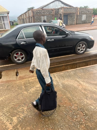 A young boy, likely a student, is seen walking down a street in Sabojibi, Nigeria, carrying a black bag. He is wearing a white shirt and dark blue trousers, typical school attire. The boy is walking past a black car, which appears to be parked on the side of the road. The street is paved with concrete and shows signs of wear. In the background, there are buildings made of concrete blocks, some with corrugated iron roofs, suggesting a residential or mixed-use area. The sky is overcast, indicating a cloudy or possibly rainy day. The overall impression is that of an ordinary day in a Nigerian town, with daily activities like commuting to or from school taking place. There is no visible text in the image.