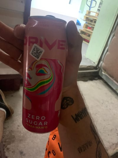 A close-up, eye-level, horizontal medium shot captures a person holding a pink aluminum can labeled "DRIVE" in the city of Kokhma, Russia. The can features a graphic of a stylized face with swirling rainbow colors and the text "ZERO SUGAR" with "ГУАВА-МАНГО" below it. A QR code is visible on the upper portion of the can. The person's hand, with visible tattoos on the forearm, is gripping the can. The background shows a dimly lit interior with a light-colored wall, a doorway, and industrial shelving with wooden pallets stacked behind. The floor is a rough, light-colored surface. The lighting is somewhat dim and uneven.