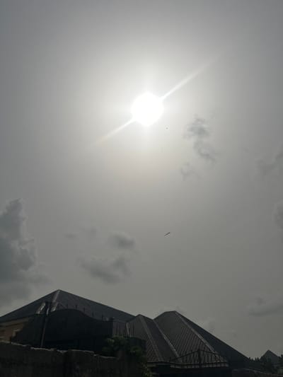 This outdoor image presents a bright, hazy sky over the city of Obinze, Nigeria. A prominent, intensely luminous sun with a distinct starburst effect occupies the upper-center to upper-left quadrant, dominating the sky. Scattered patches of light to dark grey clouds are visible, primarily in the lower-left and upper-right areas. Several dark avian figures are observed in flight against the sky, including one larger bird with outstretched wings in the middle-right. The lower third of the frame shows the dark silhouettes of multiple building rooftops, characterized by geometric, likely pyramidal or hip designs, constructed with dark-colored materials, possibly corrugated metal. A substantial dark-colored concrete or stone wall runs across the bottom of the image, with decorative metal railings visible atop sections of it and between rooflines. Sparse, dark green foliage is visible growing near the wall. No human figures or direct human activities are present.
