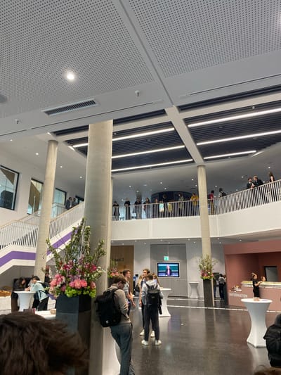 This media file captures a busy, multi-story atrium of a modern building, likely a university or public institution, seen from an elevated perspective. The interior features high ceilings with prominent linear LED lighting strips and perforated acoustic panels in light gray. Large, round concrete pillars extend vertically through the space, supporting the structure.

On the ground floor, a dark gray floor contrasts with light-colored walls. Numerous individuals are visible, casually dressed and mingling. In the mid-ground, a white "INFORMATION" desk with a white tablecloth is staffed by two people. Nearby, another person stands by a second similar white table. A large flat-screen monitor is mounted on a wall, displaying a person speaking, possibly part of a presentation or announcement. Large black planters filled with vibrant floral arrangements in pinks, greens, and white are strategically placed throughout the space, adding color. Faintly visible on a wall behind one planter is the text "Saal 2" (German for "Hall 2"). A green exit sign is also visible near the monitor.

The upper levels feature white staircases with distinctive parallel balustrades, and white railings overlooking the ground floor. More people are scattered on these upper walkways and landings, some looking down into the atrium. The architecture is clean and contemporary, with some walls featuring purple accents near the stairwells and a section of wall in a reddish-brown hue behind the information desk.

The overall scene suggests an event, such as an open day, conference, or student gathering, given the "INFORMATION" desk, the mingling crowd, and the presentation on the screen. The setting, possibly in Heidelberg, Germany, aligns with the modern institutional aesthetic. The lighting indicates it is likely daytime or an indoor event well-lit by artificial lights.