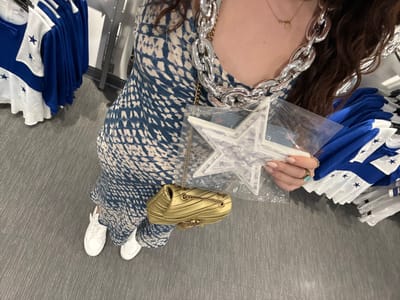 A person stands on a grey carpet, dressed in a blue and white patterned jumpsuit and white sneakers. They're surrounded by blue and white flags, holding a star-shaped item packaged in plastic. A gold purse sits atop the flags. A chunky silver necklace adorns their neck. The perspective is from above, capturing the person's attire and the surrounding merchandise.