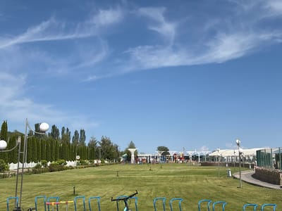 The image depicts a bright, sunny day in a park setting in Sary-Oy, Kyrgyzstan. The sky is a clear blue with wispy white clouds. The foreground features a neatly mown green lawn with several blue bicycle racks. To the left, a lamppost with two spherical lights stands tall. In the middle ground, a white picket fence lines a garden area with manicured trees and flowering bushes. Beyond this, a playground area is visible with colorful structures and canopies. A few people can be seen in the distance in the playground area, suggesting a leisurely day out. Further to the right, a curved pathway runs alongside a raised flower bed. The overall impression is one of a well-maintained recreational space, likely a tourist attraction or a public park, bathed in the warm light of midday. A speed limit sign with "20" is visible on a pole in the middle ground, indicating a controlled speed within the park.