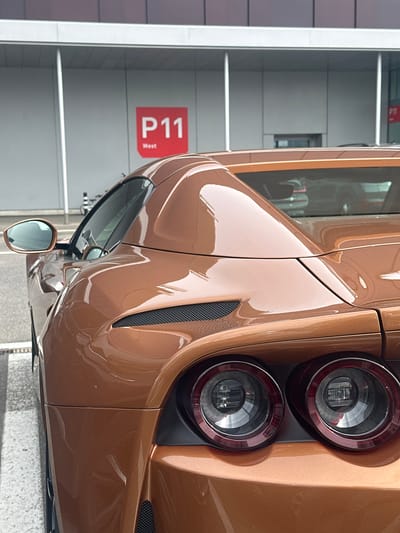 The image captures a detailed, rear three-quarter view of a modern, metallic bronze or copper-colored sports car. The car is parked indoors, likely within a clean, well-lit parking facility or showroom. The vehicle's paintwork is highly reflective, showcasing sleek, sculpted body lines and highlights from the ambient lighting.
Key features of the car include two distinct circular LED taillights on the right side of the rear, set within black housing. A black mesh grille is visible on the upper side bodywork, suggesting an air intake or vent. The driver's side mirror is prominently displayed, reflecting parts of the indoor environment. The car's overall design exudes high performance and luxury.
In the background, a light-colored, panelled wall serves as the backdrop. Affixed to this wall is a red rectangular sign with white text that reads "P11 West," indicating a specific parking level or section. The floor beneath the car appears to be a grey, textured surface, typical of a parking garage.
No people or activity are present in the image; the car is stationary. The lighting is even and diffuse, consistent with an indoor daytime setting, without direct sunlight. The image is oriented vertically, with the car appearing on its side, but is described as if viewed from a standard horizontal perspective. While the location context of Wallisellen, Switzerland is provided, there are no specific visual cues within the image itself to directly confirm this location beyond the general appearance of a modern, well-maintained facility.