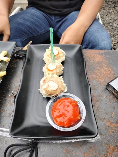 The image depicts a close-up, eye-level view of a casual outdoor or semi-outdoor dining scene in Dramaga, Indonesia, during the daytime. A person is seated, visible from the waist down, wearing a black t-shirt and blue jeans.
The main focus is on a black, rectangular, textured plate holding three pieces of siomay (steamed dumplings). Each siomay is topped with a small orange slice and a swirl of white sauce, and a green plastic mini-fork is stuck into one of them. Adjacent to the siomay, a small clear plastic cup contains a vibrant red chili sauce.
To the left of the main plate, a portion of another black plate with what appears to be french fries is visible. A set of car keys, consisting of a black remote and a silver key, rests on the table between the two plates. To the right, on the rustic, possibly metal or stone table surface, lies a partially visible pack of cigarettes. The brand names "BINTANG" and "MENTHOL" are clearly discernible on the cigarette packaging. The background is slightly blurred, showing a paved or gravel area, suggesting an informal outdoor setting. The scene captures a relaxed moment, likely a snack or meal.