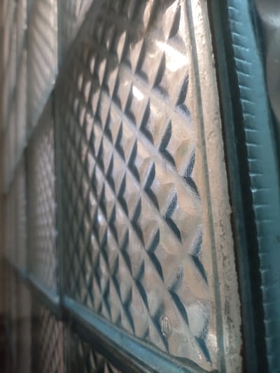 The image shows a close-up of a textured glass surface, likely a glass block or pane. The texture consists of a diamond pattern, with raised areas and recessed portions that create a play of light and shadow. The glass appears somewhat dirty or smudged, particularly along the edges and in the recessed areas of the texture. In the upper left and right corners of the image, the light is brighter, suggesting a light source is present. The color palette is muted, with shades of gray, white, and hints of blue. There are no people or discernible actions taking place. The image is an abstract representation of a textured surface. The location in the city of Stepanivka, Ukraine is not identifiable from the visual information provided in the image.
