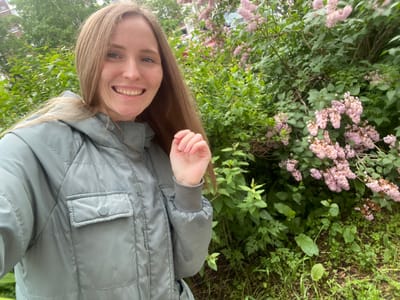 A young woman with long, blonde hair is taking a selfie in front of a blooming lilac bush. She is wearing a light grey, quilted jacket with a chest pocket. Her left hand is raised and her fingers are slightly curled. The background shows lush green foliage and clusters of pinkish-purple lilac flowers. Behind the bushes, parts of a building and a red vehicle are visible. The scene appears to be outdoors in a residential area, possibly in Yuzhno-Sakhalinsk, Russia, given the presence of lilac bushes, common in the region during bloom season.