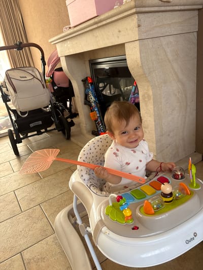 A smiling baby sits in a walker with toys. The baby is wearing a white shirt with strawberries. In front of the baby is a baby walker with colorful toys including a bee and a ladybug. The baby is holding a pink fly swatter. In the background, there is a fireplace, a stroller, and curtains. The floor is tiled. The location is the city of Veghel, Netherlands.