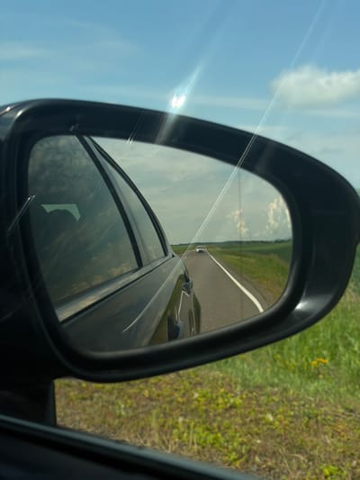 The image shows a view through a car's side mirror, reflecting a road with a white line down the center. A white car is visible in the distance on the road, which is flanked by green fields under a blue sky with scattered clouds. Sunlight is creating lens flare in the upper portion of the image. The car exterior is dark and blurred in the foreground, and some of the car's interior can be seen through the side window. There is no indication of Krasnaya, Russia.